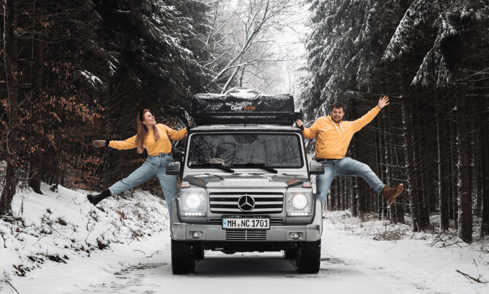  Pareja con ropa a juego junto a su coche con tienda de techo Campwerk en una carretera nevada en el bosque