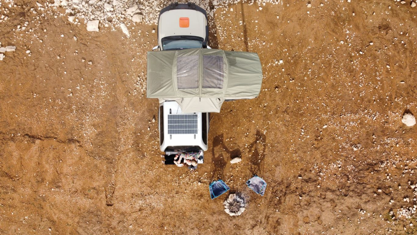 Tienda de techo Adventure de Campwerk desde la perspectiva de un pájaro. Una pareja está sentada en la parte trasera del coche, junto a ellos hay sillas de camping y una hoguera 